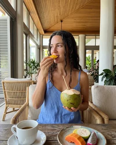 Casual smartphone photo of a woman with braided dark hair holding a coconut drink and mango at an upscale tropical resort lounge, warm honey-toned wood