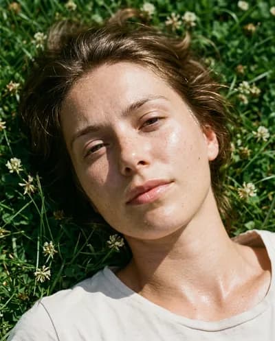 Raw photorealistic portrait lying on grass in harsh sunlight, dewy skin with visible freckles, white wildflowers around