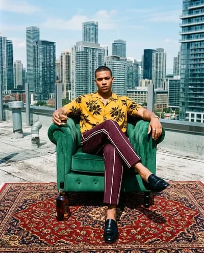 Homme en chemise florale moutarde assis dans un fauteuil émeraude sur un toit avec de l'alcool, un tapis persan et une ligne d'horizon de la ville derrière