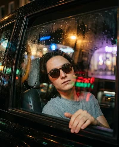 Shot through rain-spattered taxi window, subject in dove-grey t-shirt and tortoiseshell sunglasses, neon bokeh cityscape, teal-amber grade