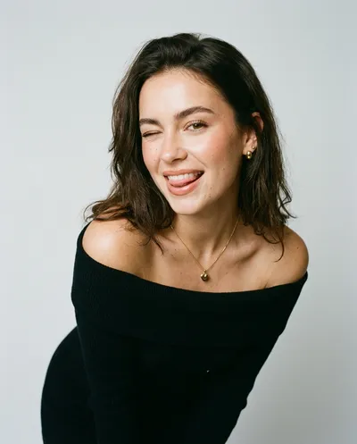 Studio portrait of a young woman winking playfully with tongue slightly out, wearing a black off-shoulder dress and gold heart pendant, clean minimal background