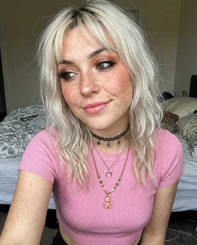 Selfie de primer plano de una mujer pecosa con pelo rubio platino, maquillaje brillante y collares en capas con luz íntima de dormitorio