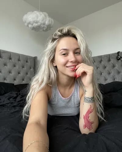 Foto casual de smartphone de una mujer rubia platino con tatuaje de pez koi acostada en una cama con sábanas negras mirando a la cámara