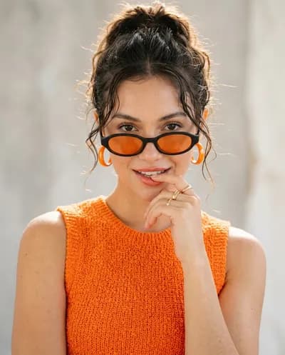 Retrato editorial de moda de una mujer con accesorios de color mandarina vibrante con una pose juguetona de dedo a labios y un cálido halo retroiluminado.