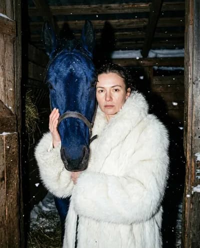 Nighttime flash portrait with woman in white fur coat beside a surreal cobalt-blue horse in a wooden stable, frozen snowflakes in the air