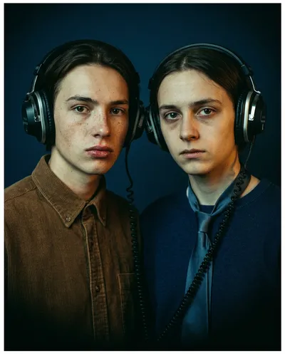 Dual portrait of two young people in vintage headphones, moody indigo background, serious direct gaze, retro film aesthetic