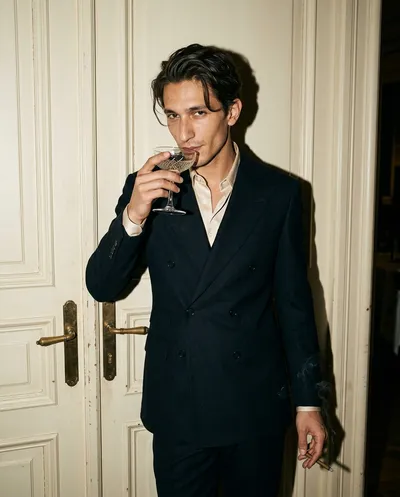 Portrait flash d'un homme en costume bleu nuit appuyé contre des portes crème, verre coupe cristal aux lèvres, fumée de cigarillo s'élevant