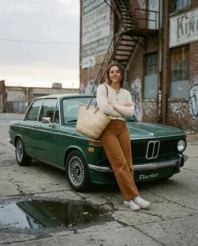 Candid full-body street photography, urban warehouse district at dusk. A Forest Green 1974 BMW 2002 Turbo, front three-quarter view, chrome...
