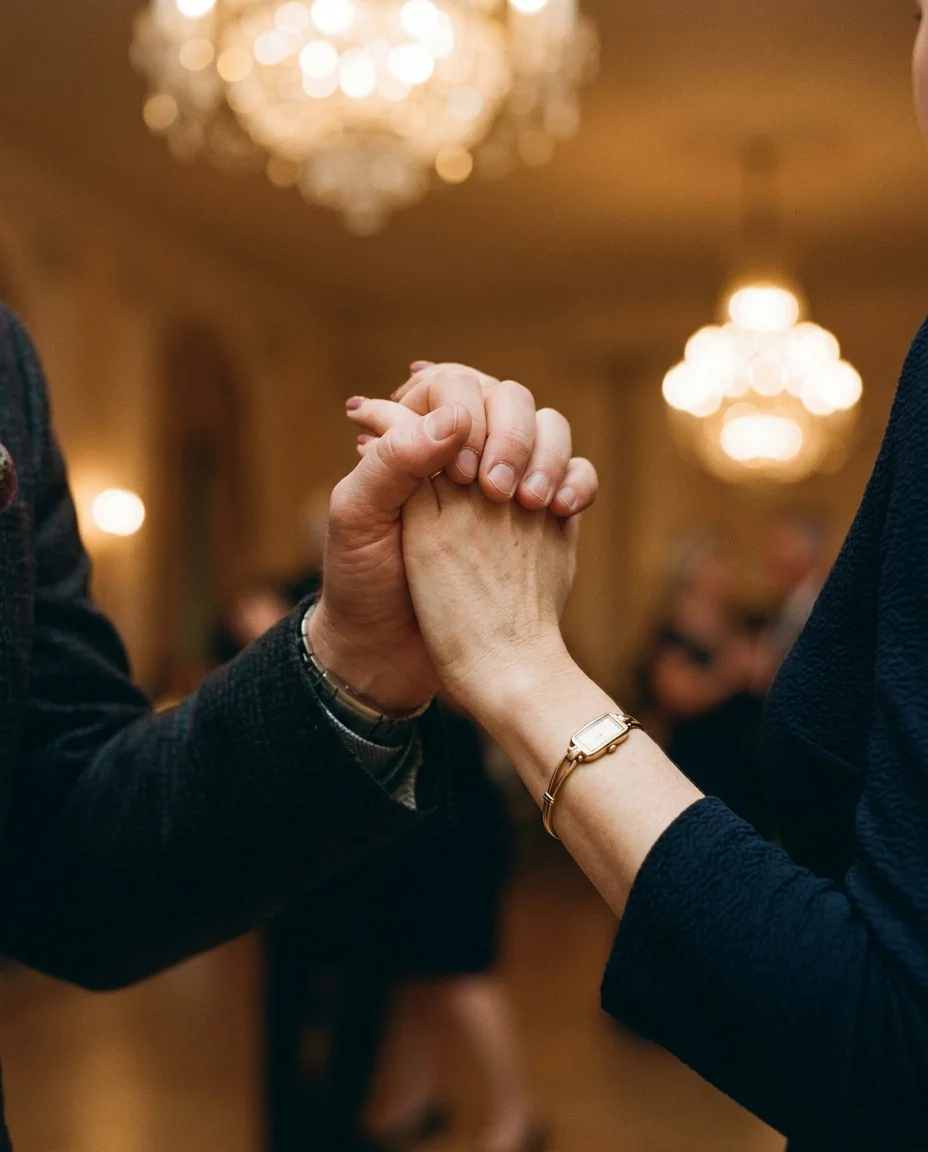 لقطة مقربة لأيدي زوجين متشابكة أثناء الرقص، كم قميصه الساتاني العاجي يلتقي بكم فستانها المخملي الكحلي، وخاتم ختم ذهبي يلتقط ضوء الشموع.