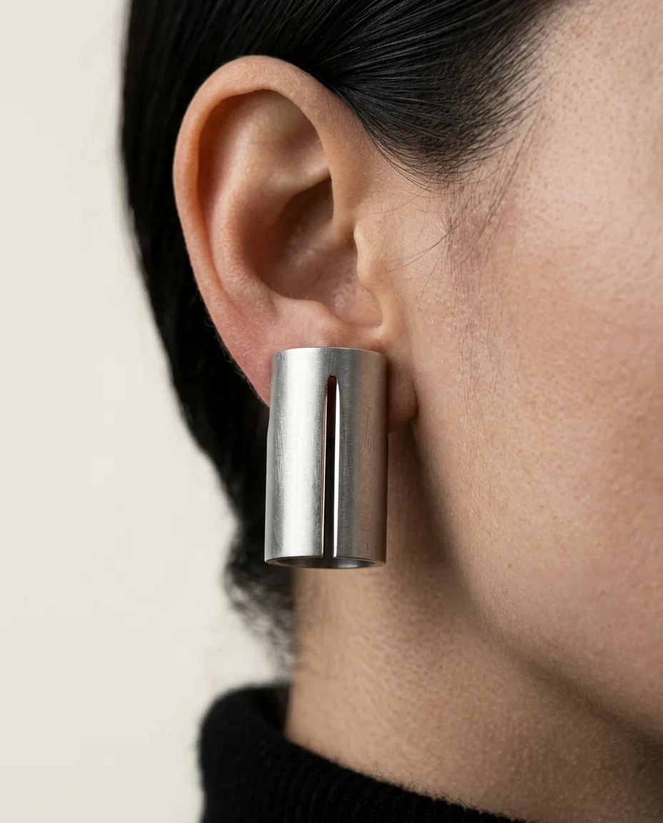 Extreme macro close-up of an ultra-minimal matte silver cylindrical cuff earring on the model's left ear. The frame is filled...