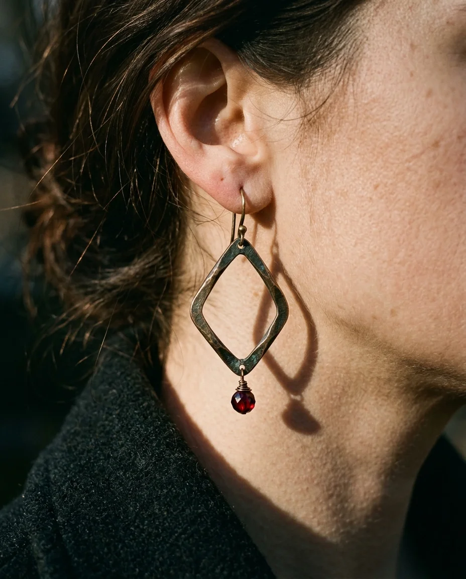Extreme close-up editorial photograph focusing on the left ear and the angular geometric drop earring in oxidized bronze. The open...