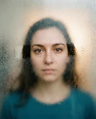 صورة فوتوغرافية فائقة الواقعية لصورة شخصية امرأة تُرى من خلال لوح زجاجي سميك ضبابي مصنفر، مع وجود جزء أفقي صغير فقط...