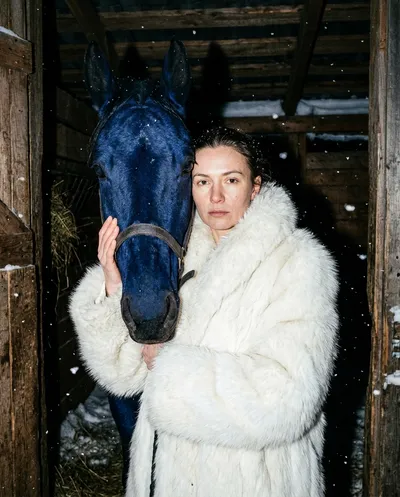 Nighttime flash portrait with woman in white fur coat beside a surreal cobalt-blue horse in a wooden stable, frozen snowflakes in the air